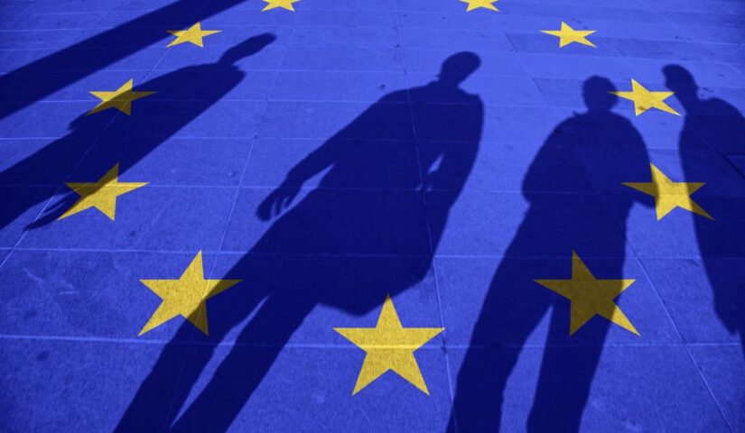 Na chodniku cienie kilku osób. Na chodnik nałożony jest niebieski filtr z gwiazdkami Unii Europejskiej