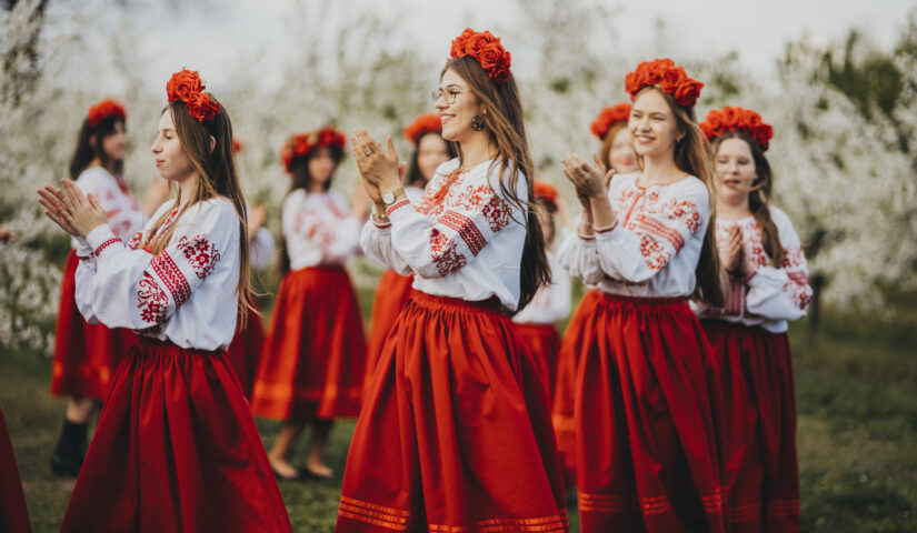 Tancerki w strojach ludowych tańczą w kole. W tle widoczne kwitnące drzewa owocowe.